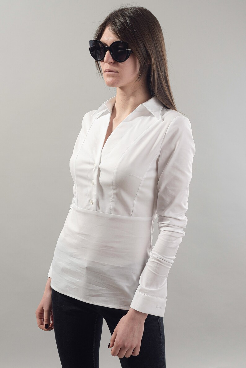 Puede incluir: Una camisa blanca de manga larga con botones y cuello en V. La camisa tiene una cintura ajustada y est&aacute; metida en un pantal&oacute;n negro.