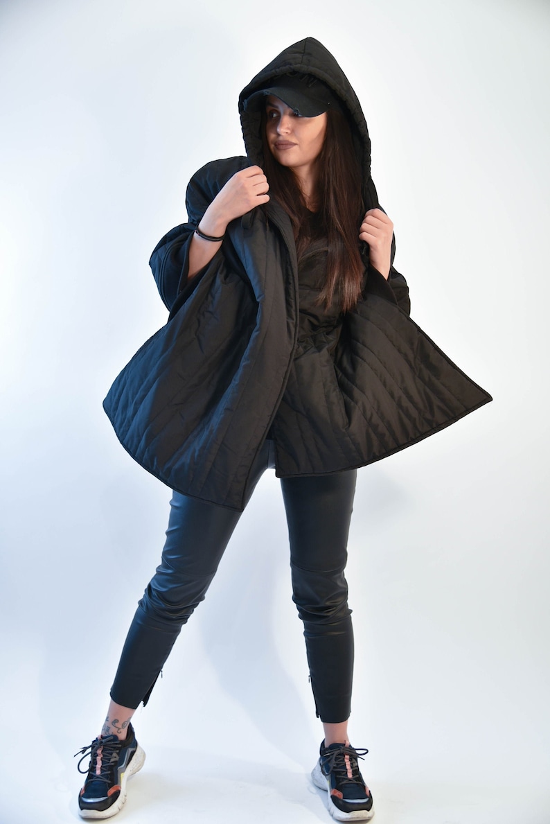 May include: A black quilted jacket with a hood and a black baseball cap. The jacket is open and the person is wearing black pants and black sneakers.