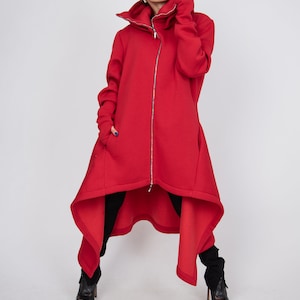 Könnte beinhalten: Eine leuchtend rote Langarmjacke mit hohem Kragen, einem durchgehenden silbernen Reißverschluss und einem asymmetrischen Saum, der hinten länger ist. Die Jacke verfügt über lange Ärmel mit Daumenlöchern und tiefen Seitentaschen über schwarzen Hosen.