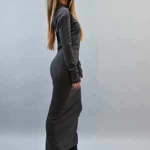 May include: A long, dark gray dress with long sleeves and a high neckline. The dress is form-fitting and reaches the floor. The person is wearing black open-toe high heels. The background is a solid light gray.