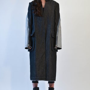 Op de afbeelding: Een lange zwarte denim jas met ritsdetails op de mouwen. De jas heeft een kraag en wordt gedragen door iemand met een zonnebril.
