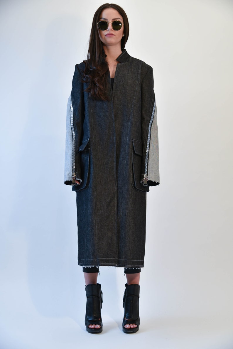 Op de afbeelding: Een lange zwarte denim jas met ritsdetails op de mouwen. De jas heeft een kraag en wordt gedragen door iemand met een zonnebril.