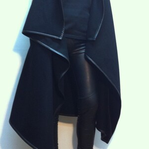 May include: A black sleeveless vest with a draped open front and leather-look trim, worn over a black tank top and black leather-look leggings. The outfit is completed with black platform peep-toe wedge heels. The text Flo Atelier is visible.