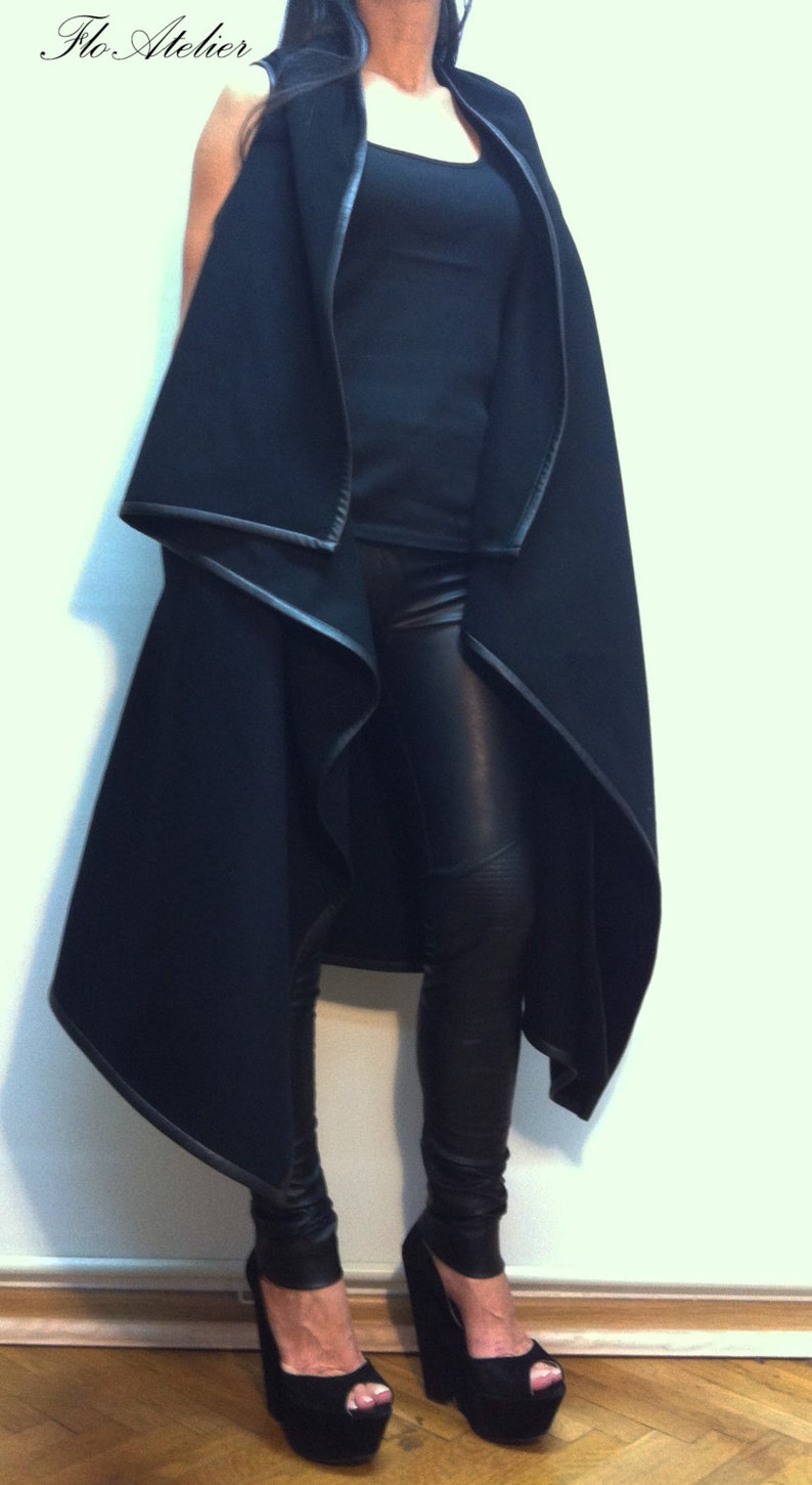 May include: A black sleeveless vest with a draped open front and leather-look trim, worn over a black tank top and black leather-look leggings. The outfit is completed with black platform peep-toe wedge heels. The text Flo Atelier is visible.