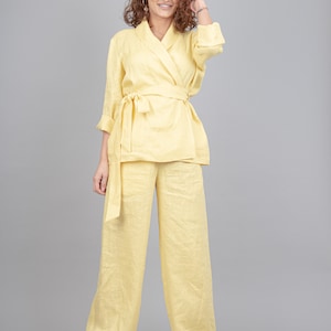 May include: A woman wearing a yellow linen pantsuit with a long-sleeved, wrap-style top and a matching pair of wide-leg trousers. She is smiling and has curly brown hair.
