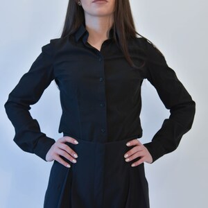 May include: A woman wearing a black long-sleeved shirt and black pants with a high waist. She is standing with her hands on her hips.
