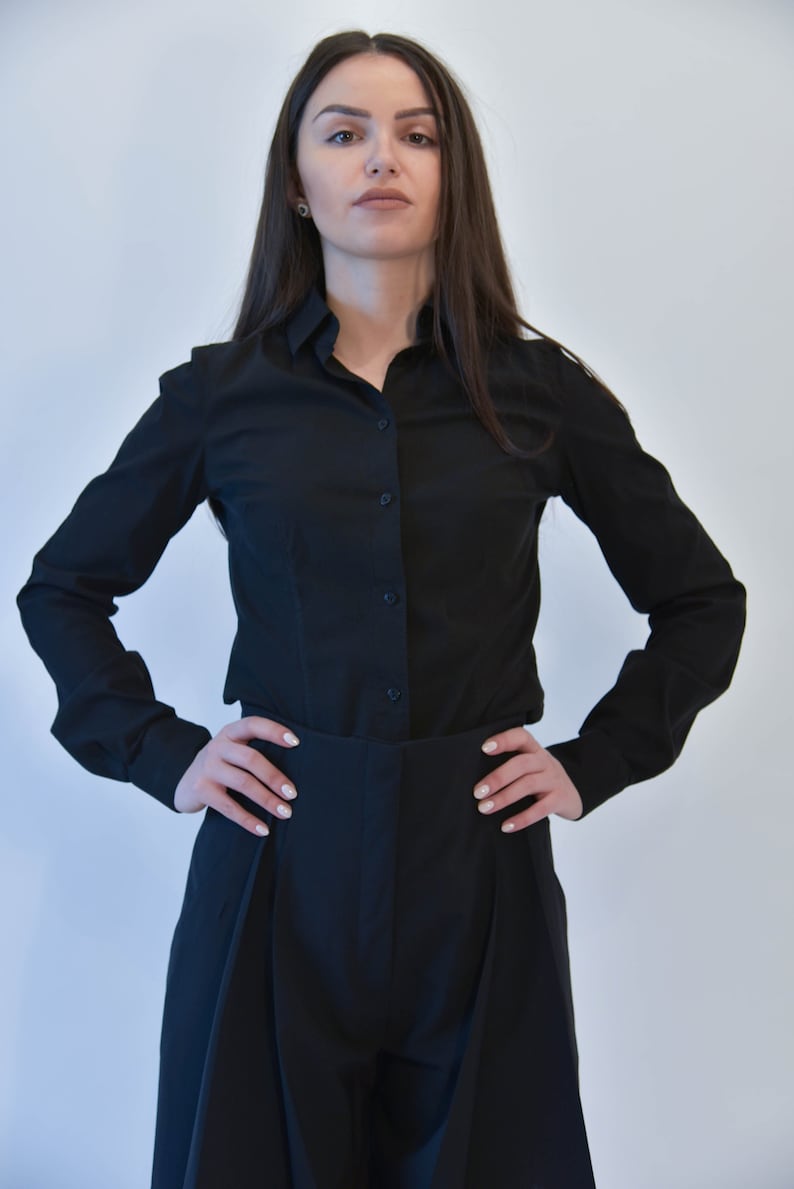 May include: A woman wearing a black long-sleeved shirt and black pants with a high waist. She is standing with her hands on her hips.