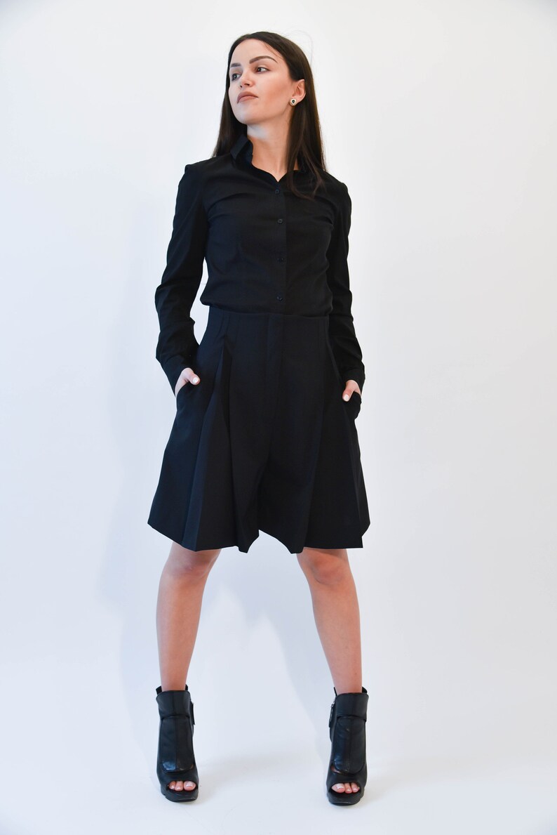 May include: A woman wearing a black long-sleeved shirt and black shorts with a pleated design. She is wearing black heeled ankle boots.