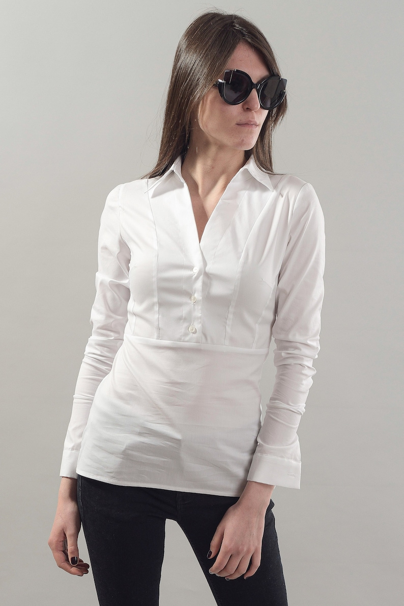 Puede incluir: Una camisa blanca de manga larga con botones, cuello en V y cintura ajustada. La camisa la lleva puesta una persona que lleva pantalones negros y gafas de sol.