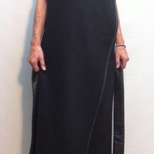 May include: A long, sleeveless black wrap dress featuring a wide, folded collar and dark leather-like trim along the edges. The garment has a straight, floor-length silhouette with long, hanging fabric panels at the sides and a clean finish.
