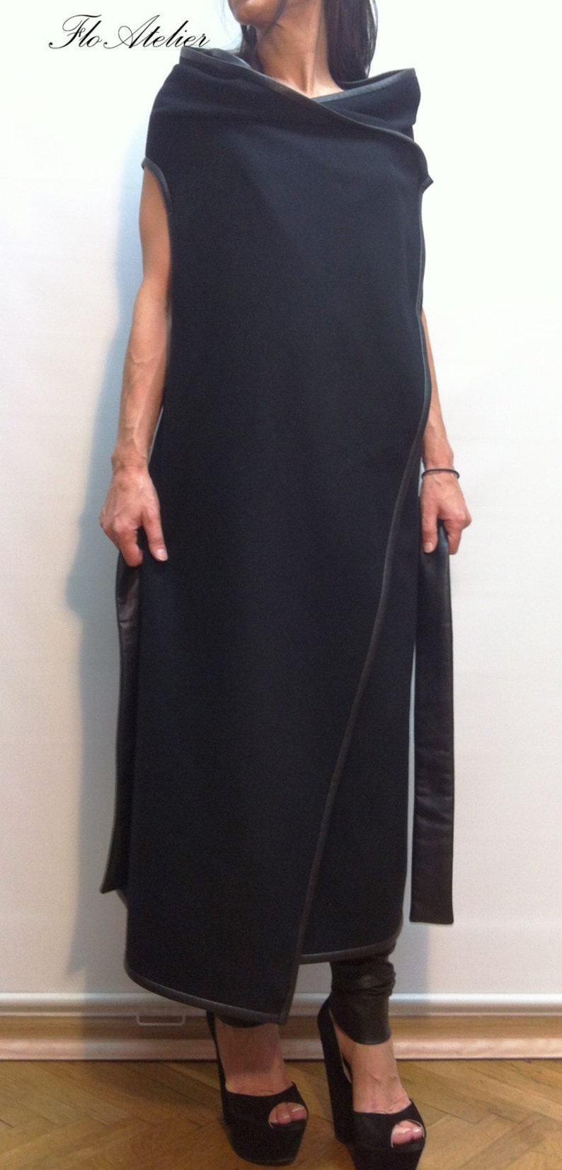 May include: A long, sleeveless black wrap dress featuring a wide, folded collar and dark leather-like trim along the edges. The garment has a straight, floor-length silhouette with long, hanging fabric panels at the sides and a clean finish.