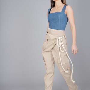 May include: A woman wearing a denim blue top with a square neckline and beige cargo pants. The pants have a wrap-around design with a white rope tie. Beige woven sandals complete the outfit.