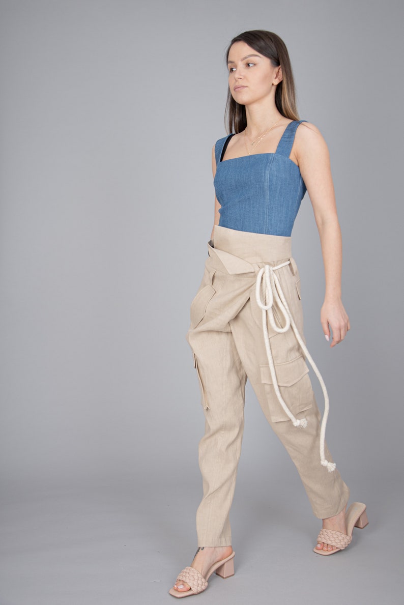 May include: A woman wearing a denim blue top with a square neckline and beige cargo pants. The pants have a wrap-around design with a white rope tie. Beige woven sandals complete the outfit.