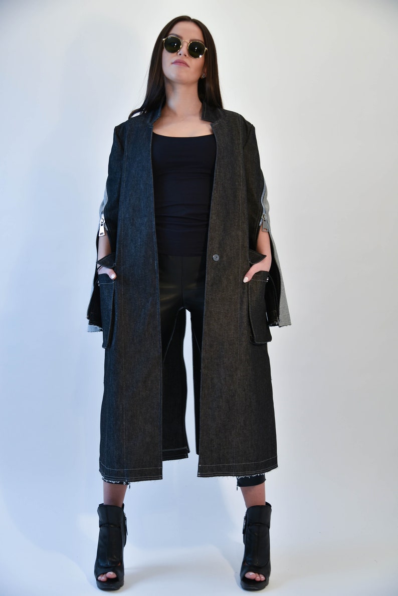 Op de afbeelding: Een lange zwarte denim jas met ritsdetails en een enkele knoopsluiting. De jas heeft een losse pasvorm en wordt gedragen over een zwart topje en een zwarte broek.