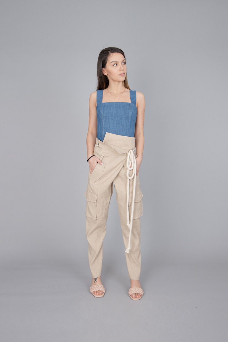 May include: A denim blue top with wide straps and a square neckline paired with beige cargo pants. The pants feature large pockets and a wrap-around waist with a white rope tie. The model is wearing light pink sandals.
