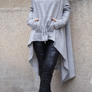 May include: A long, gray, hooded sweatshirt with a cowl neck and a high-low hemline. The sweatshirt has a drawstring hood and two pockets. The model is wearing black leggings with a floral pattern and black boots.