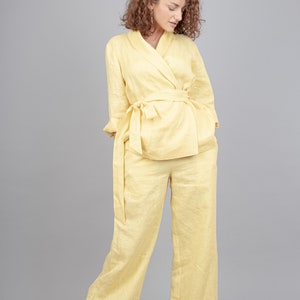 May include: A woman wearing a yellow linen trouser suit with a matching belt. The trouser suit has a relaxed fit and wide legs. The woman is wearing white trainers.