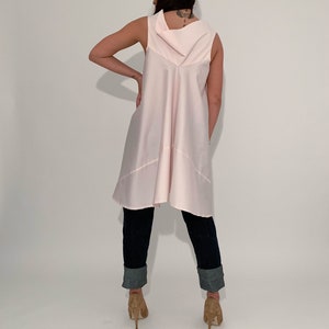 May include: A pink sleeveless tunic top with a cowl neck and a high-low hemline. The tunic is worn with dark blue jeans that have been rolled up at the ankles.