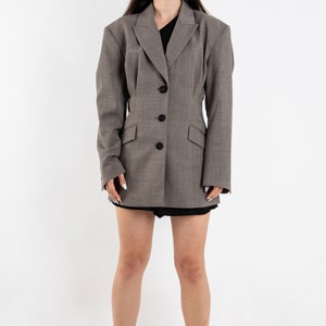 May include: A gray blazer with a fitted waist and a black button closure. The blazer has a slightly oversized fit and long sleeves. The model is wearing white sneakers.