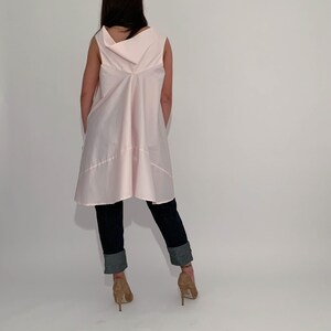 May include: A pink sleeveless tunic top with a cowl neck and a flowing, asymmetrical hemline. The top is worn with dark blue jeans and brown heeled shoes.