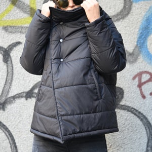 May include: A black puffer jacket with a high collar and a button closure. The jacket has a quilted design and is worn by a person wearing a black cap and sunglasses.