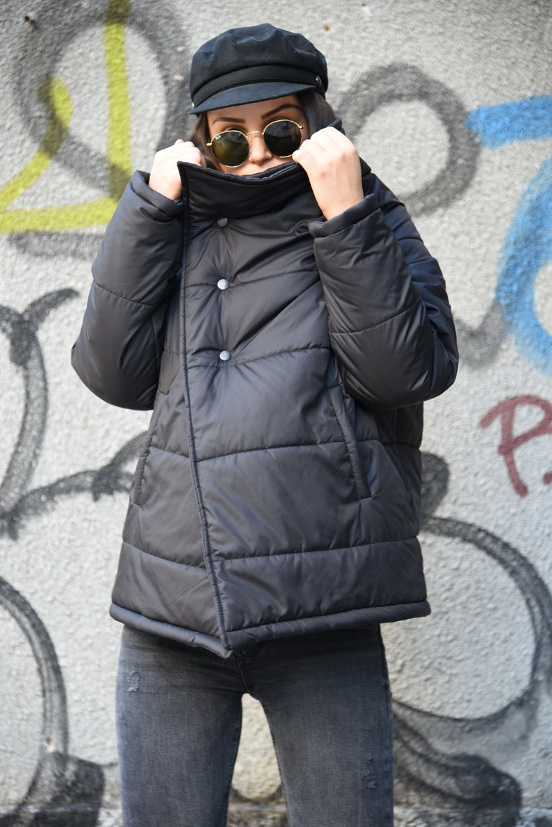 May include: A black puffer jacket with a high collar and a button closure. The jacket has a quilted design and is worn by a person wearing a black cap and sunglasses.