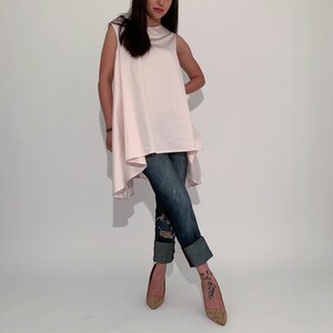 May include: A woman is wearing a light pink sleeveless top with a high low hemline and blue jeans. She is wearing brown heeled shoes.