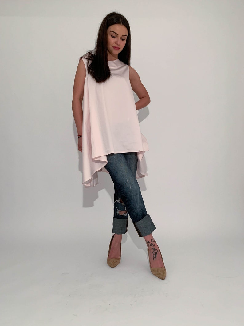 May include: A woman is wearing a light pink sleeveless top with a high low hemline and blue jeans. She is wearing brown heeled shoes.