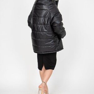 May include: A black puffer jacket with a high collar and a shiny finish. The jacket is worn by a person with long dark hair and is standing on a white background.