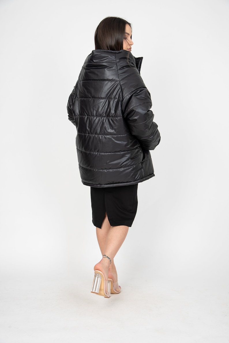 May include: A black puffer jacket with a high collar and a shiny finish. The jacket is worn by a person with long dark hair and is standing on a white background.