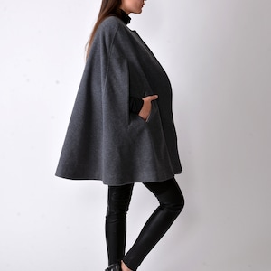 May include: A gray wool cape with a long, flowing silhouette. The cape has a high neckline and a single pocket on the left side. The model is wearing black leggings and black shoes.