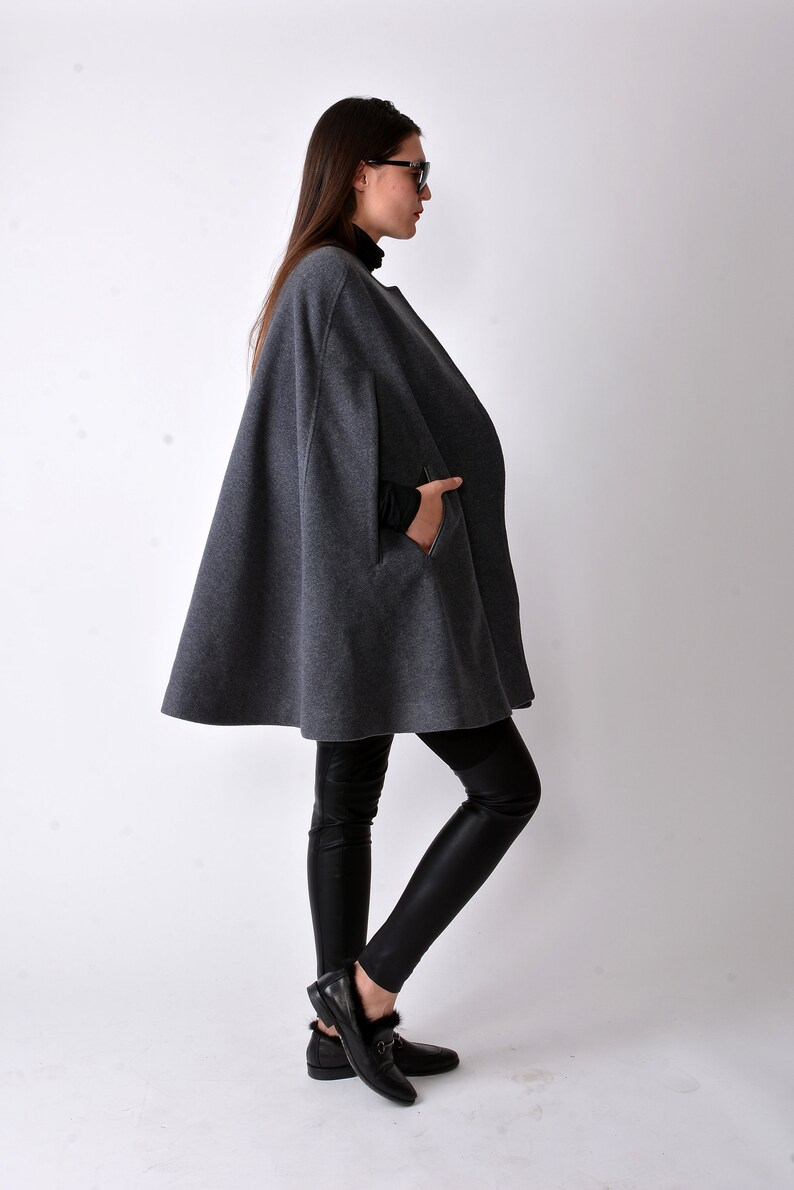 May include: A gray wool cape with a long, flowing silhouette. The cape has a high neckline and a single pocket on the left side. The model is wearing black leggings and black shoes.