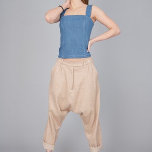 May include: A woman wearing a light blue denim tank top and beige linen trousers with a loose, wide-leg fit. She is wearing light beige sandals.
