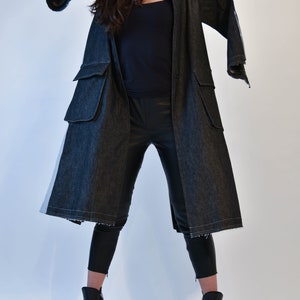 Op de afbeelding: Een lange zwarte denim jas met een witte streep aan de zijkant. De jas heeft een losse pasvorm en wordt gesloten met knopen. Het model draagt een zwarte broek en zwarte laarzen met hak.