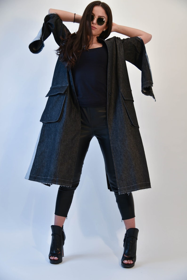 Op de afbeelding: Een lange zwarte denim jas met een witte streep aan de zijkant. De jas heeft een losse pasvorm en wordt gesloten met knopen. Het model draagt een zwarte broek en zwarte laarzen met hak.