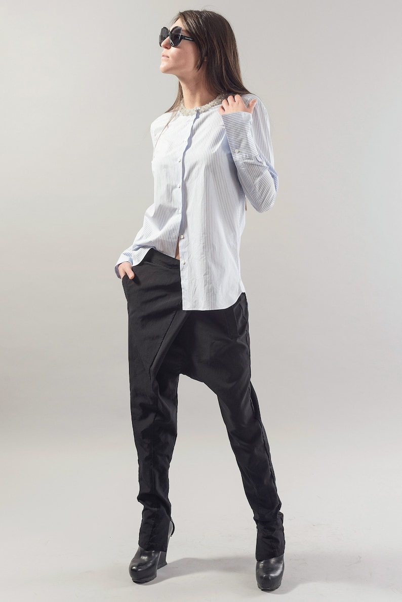 May include: A woman wearing a light blue striped button-down shirt with a pearl necklace and black trousers. She is wearing black sunglasses and has her hair pulled back.