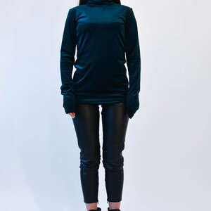 May include: A woman wearing a teal velvet turtleneck sweater and black pants with black heeled boots.