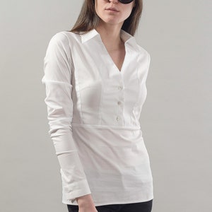 Puede incluir: Una camisa blanca de manga larga con botones, cuello en V y cintura ajustada. La camisa tiene un cierre de bot&oacute;n &uacute;nico en la parte superior y tres botones en la parte delantera. La modelo lleva pantalones negros y gafas de sol negras de ojo de gato.