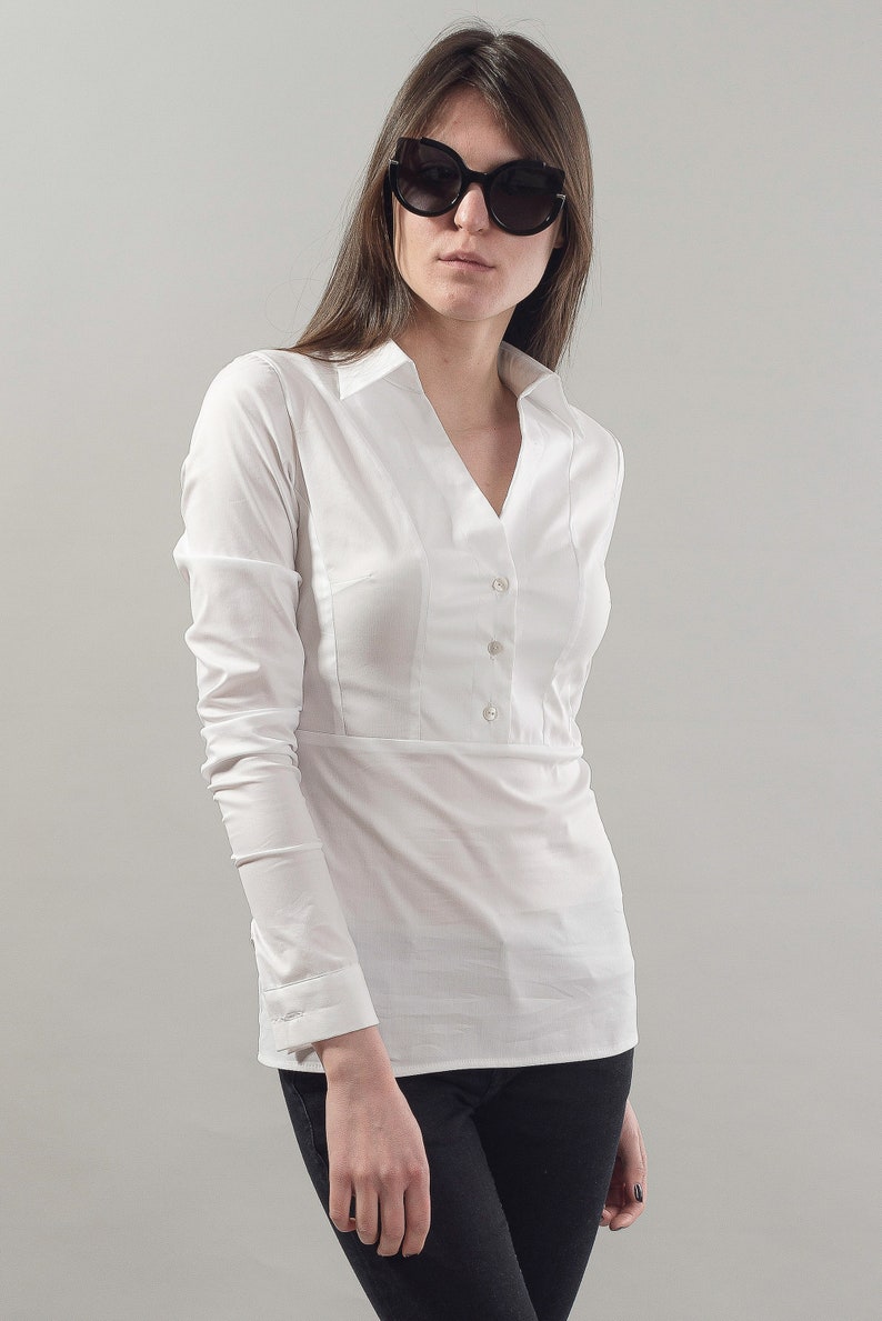 Puede incluir: Una camisa blanca de manga larga con botones, cuello en V y cintura ajustada. La camisa tiene un cierre de bot&oacute;n &uacute;nico en la parte superior y tres botones en la parte delantera. La modelo lleva pantalones negros y gafas de sol negras de ojo de gato.
