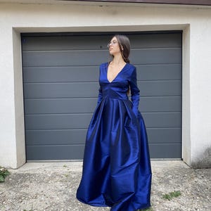 May include: A floor-length, deep blue gown featuring long sleeves, a deep V-neckline, and a full, pleated skirt. The fabric has a subtle sheen, and the dress includes integrated side pockets, creating a structured silhouette with a fitted bodice.