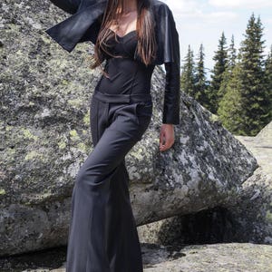 May include: A woman wearing a black leather jacket, a black top, and black wide-leg pants. She is standing on a rocky surface and looking to the right. She is wearing sunglasses.