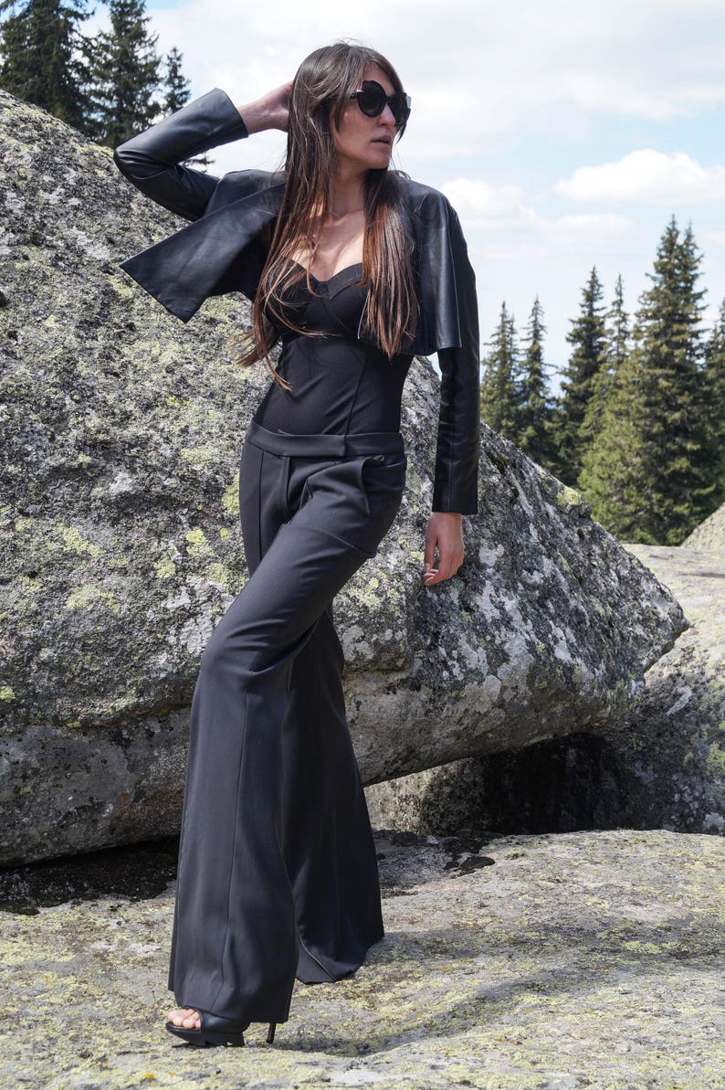 May include: A woman wearing a black leather jacket, a black top, and black wide-leg pants. She is standing on a rocky surface and looking to the right. She is wearing sunglasses.
