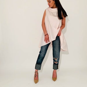 May include: A woman wearing a light pink sleeveless top with a high neckline and a long, flowing hem. She is wearing blue jeans with rolled up cuffs and brown heeled shoes.
