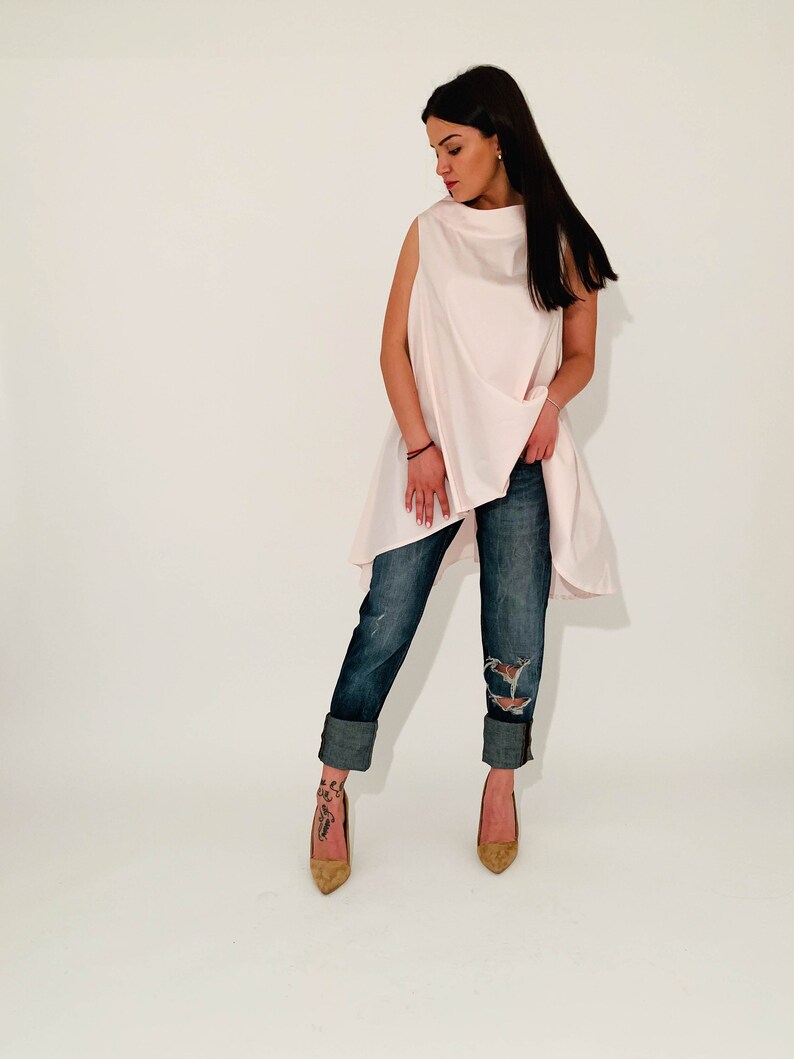 May include: A woman wearing a light pink sleeveless top with a high neckline and a long, flowing hem. She is wearing blue jeans with rolled up cuffs and brown heeled shoes.