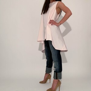 May include: A woman wearing a light pink, sleeveless, asymmetrical top with a high-low hemline. She is also wearing blue jeans with the cuffs rolled up and brown high-heeled shoes.