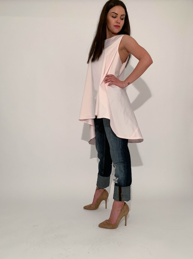May include: A woman wearing a light pink, sleeveless, asymmetrical top with a high-low hemline. She is also wearing blue jeans with the cuffs rolled up and brown high-heeled shoes.