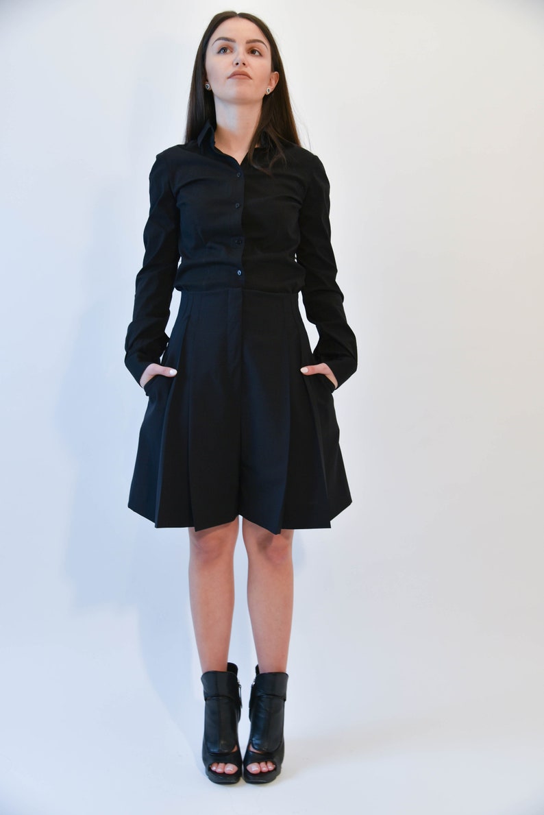 May include: A woman wearing a black button-down shirt and a black pleated skirt. She is wearing black heeled ankle boots with open toes.