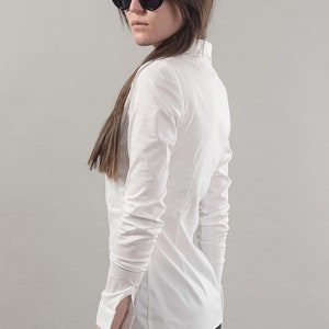 Puede incluir: Una camisa blanca de manga larga con botones y cuello. La camisa est&aacute; metida en unos vaqueros negros. La persona lleva gafas de sol negras.