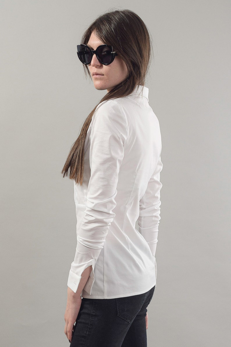Puede incluir: Una camisa blanca de manga larga con botones y cuello. La camisa est&aacute; metida en unos vaqueros negros. La persona lleva gafas de sol negras.