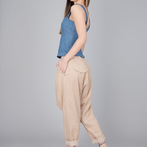 May include: A woman wearing a blue denim tank top and beige linen trousers. She is wearing beige heeled sandals and has her hair down.